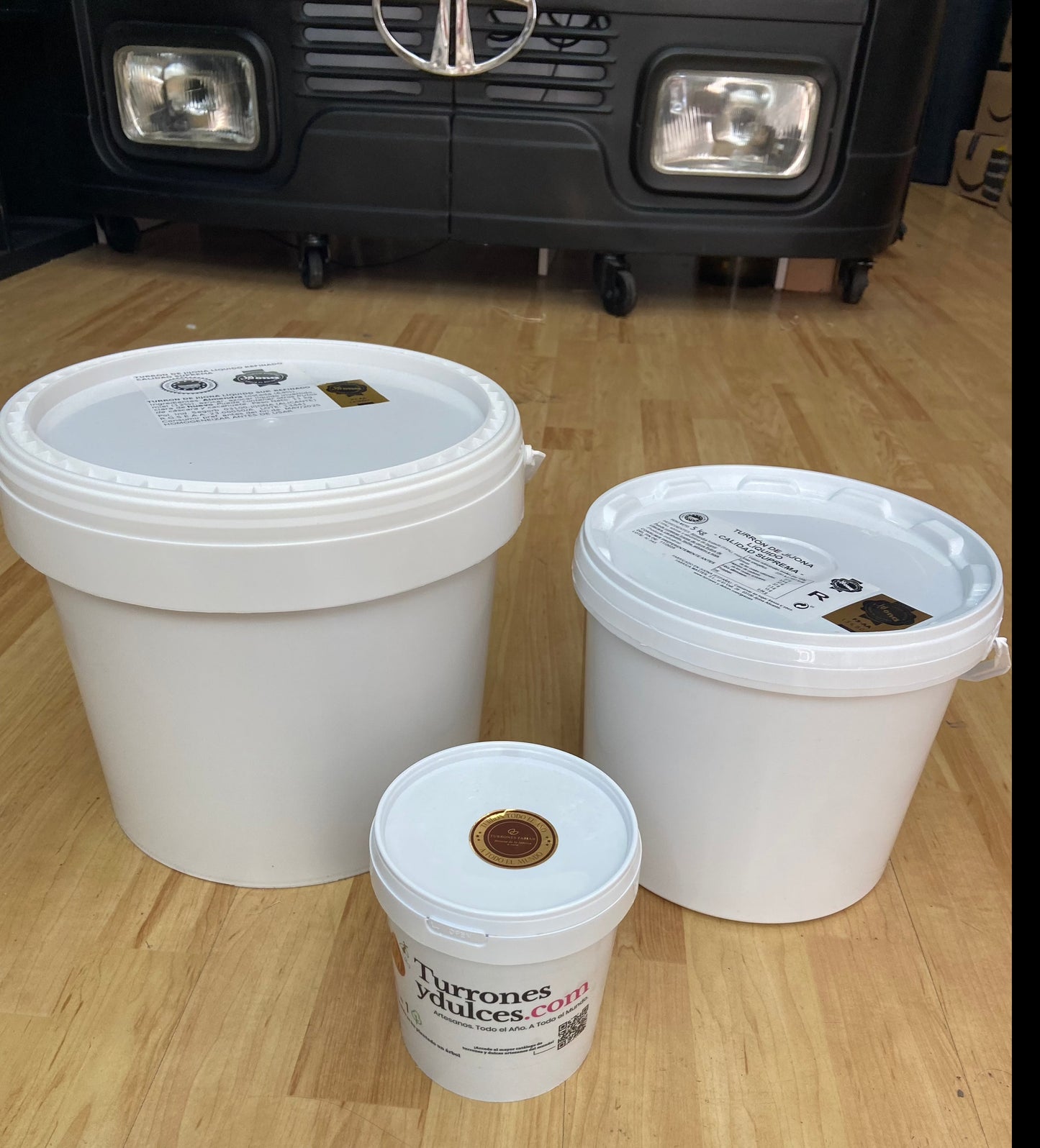 Tres tarrinas blancas (1kg, 5kg, 11kg) de Turrón líquido en pasta blando Jijona de ChocolateSI para helado y postres reposan en un suelo de madera ante un carro negro, ideal para postres y con la etiqueta "Turronesyducles.com".