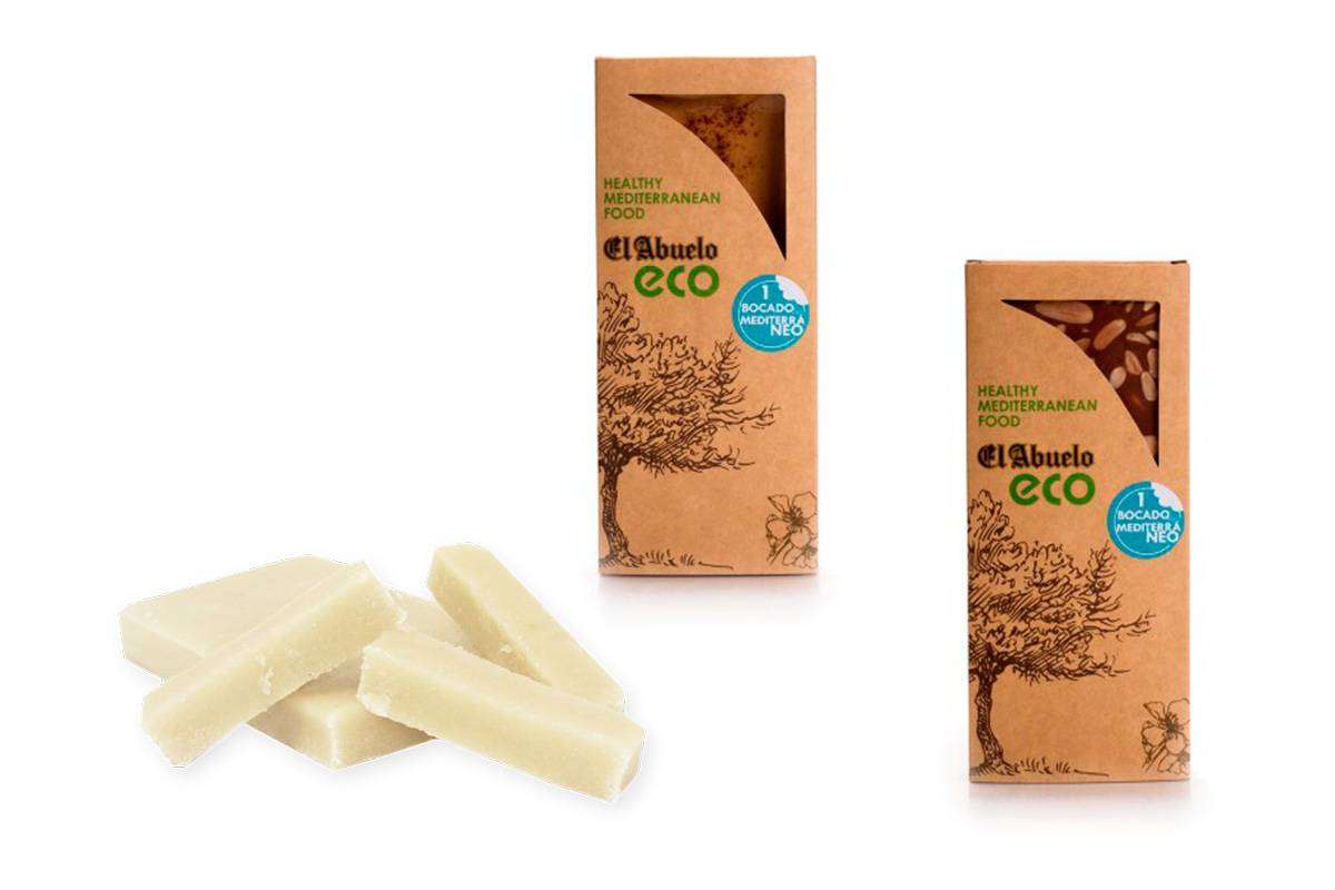 Dos cajas de "Lote de turrones para veganos, y ecológicos" de Jijona S.A., con la ilustración de un árbol, se muestran junto a varias rodajas de caramelo blanco de turrón ecológico.