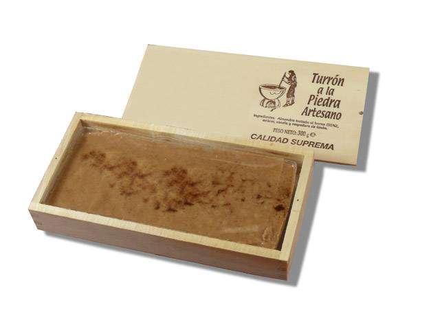 Una caja de madera con la tapa parcialmente abierta revela un bloque rectangular de "Turrón apto para veganos a la Piedra Artesano en tableta de 300g" de ChocolateSI, capturando la rica esencia de la tradición española en cada pieza.